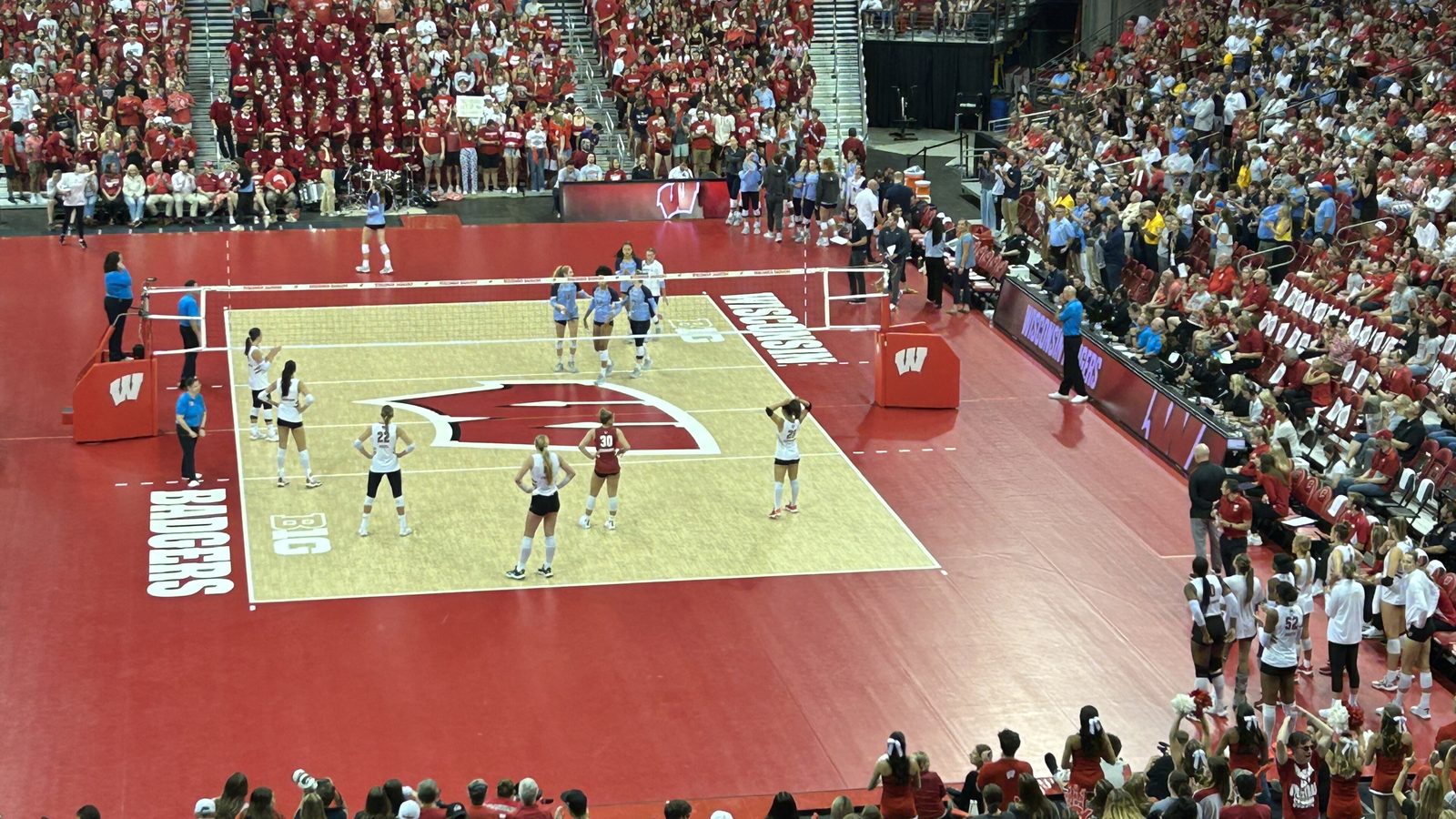 Elevated view of a women's college volleyball match at the Kohl Center, with players set in rotation on a light wood court framed by red Wisconsin branding and a packed student section behind the far end line.