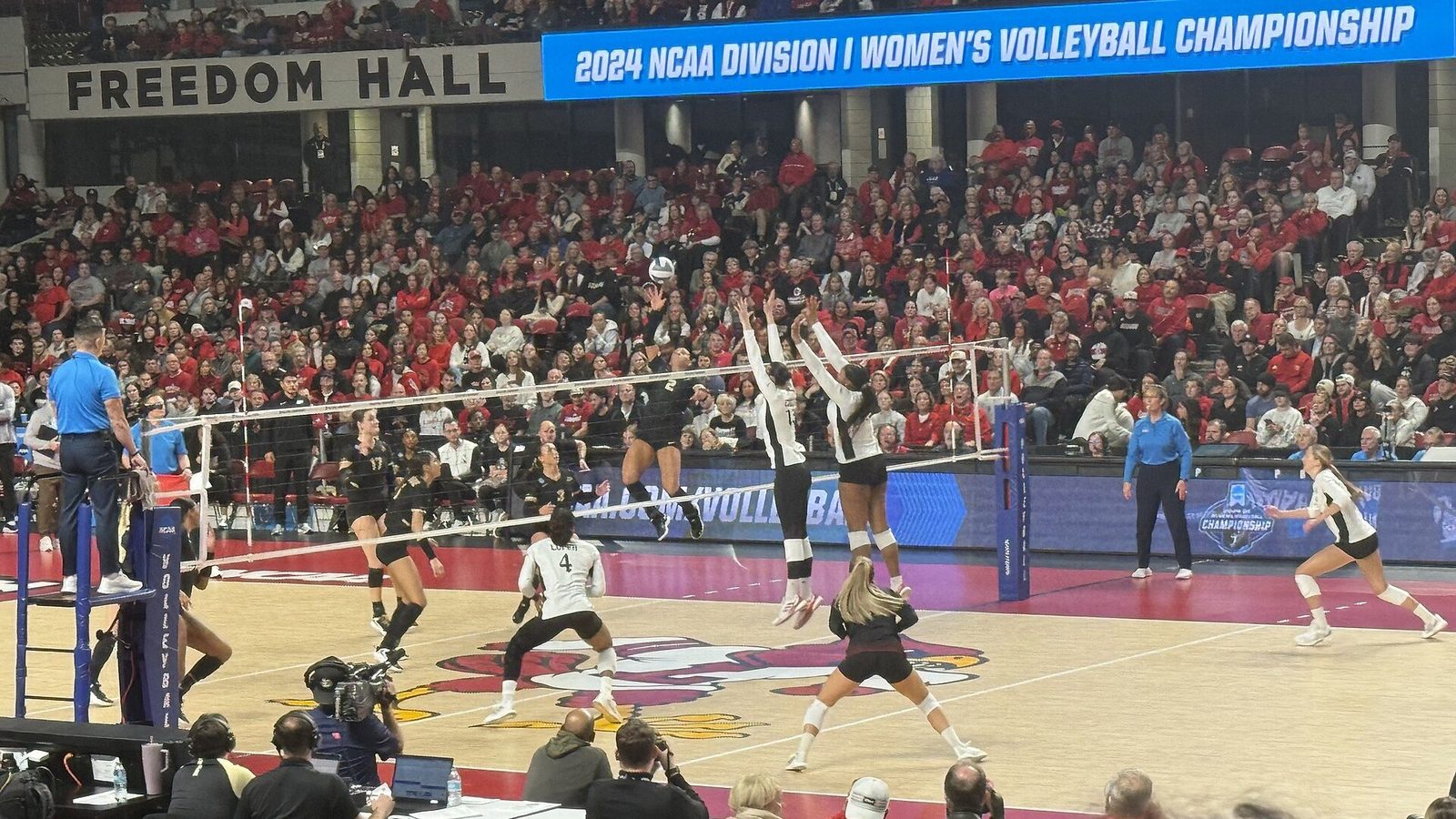 A Purdue outside hitter in black rises to attack as two Louisville blockers in white leap with arms high above the net, in front of a packed red crowd under a banner reading 2024 NCAA Division I Women's Volleyball Championship.