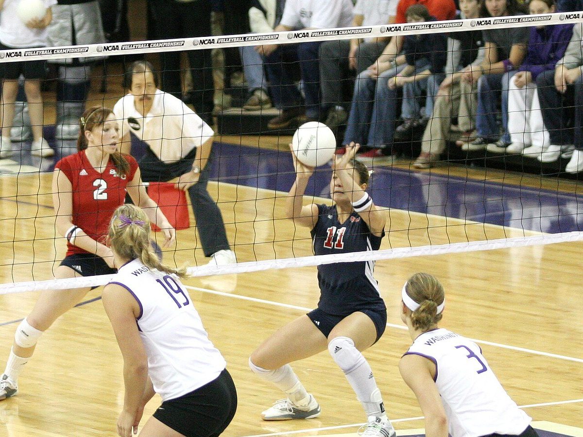 In an NCAA championship match, a player in a navy jersey number 11 crouches low and plays the ball with both hands above her face, while three opponents in white watch from the near side of the net.