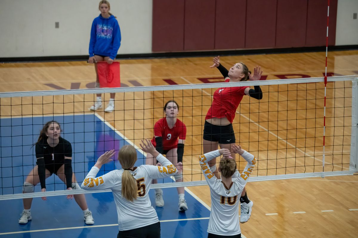 A women's indoor volleyball rally at the net: a player in red rises to attack while two players in white jump to block, and a teammate in a black jersey crouches low in a ready stance.
