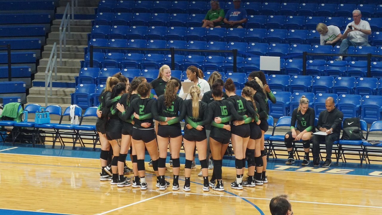 North Texas women's volleyball players in black jerseys with green sleeves huddle arm in arm around a staff member holding a clipboard at College Park Center, with mostly empty blue seats behind them.