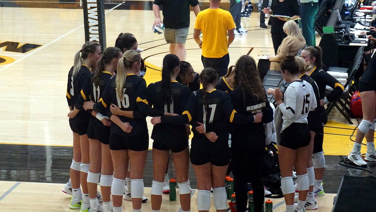 Milwaukee women's volleyball players in black and gold stand arm in arm in a timeout huddle at the Klotsche Center while staff members, one in a yellow shirt and one with a clipboard, stand nearby.