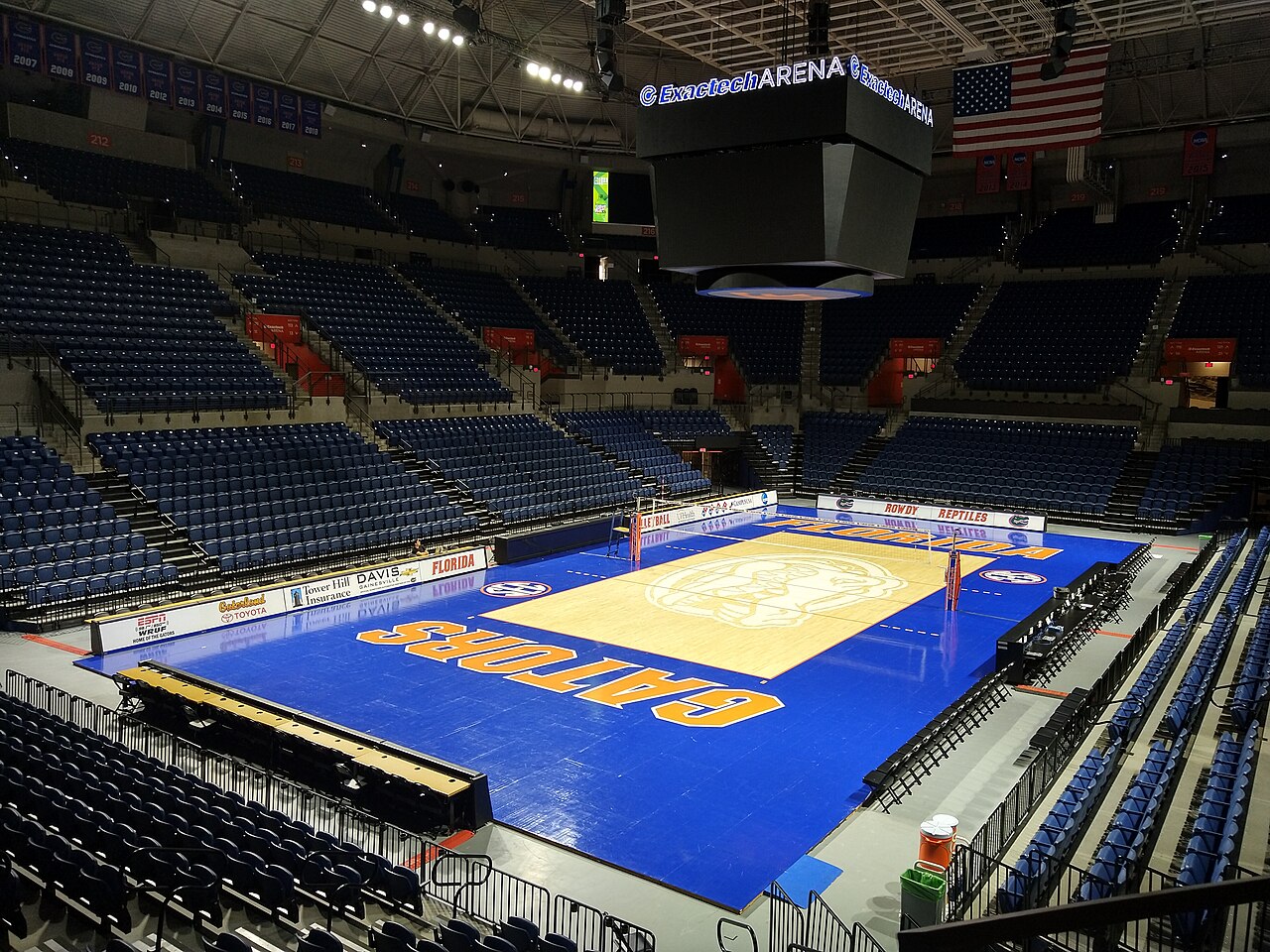 An empty Exactech Arena set up for volleyball, with a blue floor painted GATORS in orange, a wood-colored center court with the Gator logo, and rows of blue seats rising on every side.