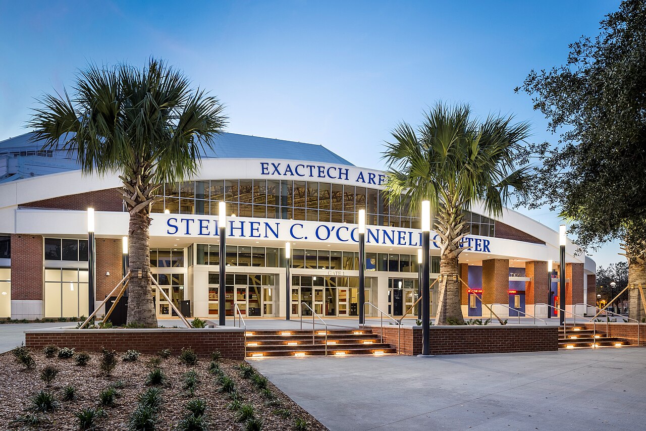 The front entrance of the Stephen C. O'Connell Center at dusk, with Exactech Arena and the building's name in blue letters above glass doors, framed by two palm trees.