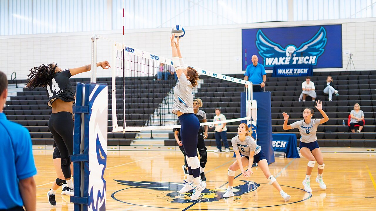 A Wake Tech Community College middle blocker reaches above the tape to block an opposing hitter's swing while her teammates cover in a bright gym with a large Wake Tech eagle banner on the far wall.
