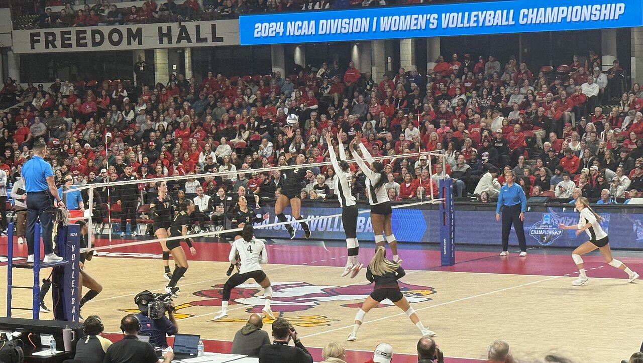 A women's volleyball match at Freedom Hall during the 2024 NCAA Division I Women's Volleyball Championship. Two Louisville blockers in white jump at the net against a Purdue attacker in black, in front of a crowd dressed mostly in red.