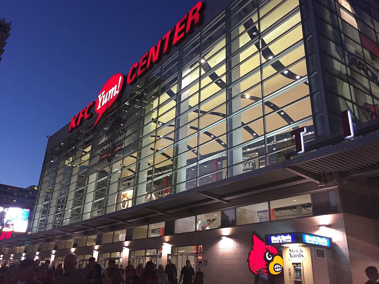 The glass facade of the KFC Yum! Center in downtown Louisville at dusk, lit from inside, with a red KFC Yum! Center sign and a large Louisville Cardinal bird logo near the entrance as fans walk past.