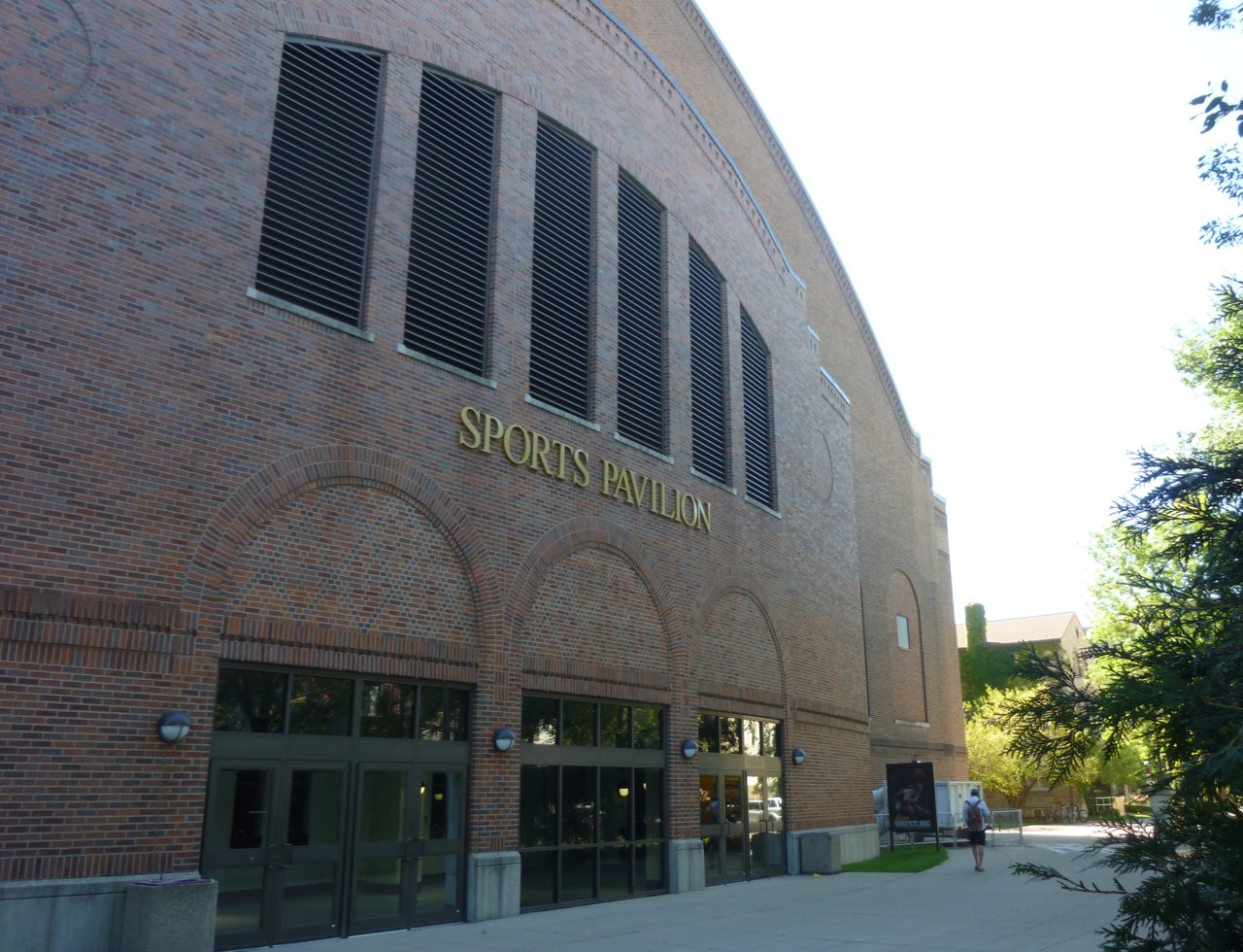 The red-brick arched facade of the University of Minnesota's Sports Pavilion, with tall slatted windows and gold letters reading Sports Pavilion above three arched glass entrances, on a sunny day.