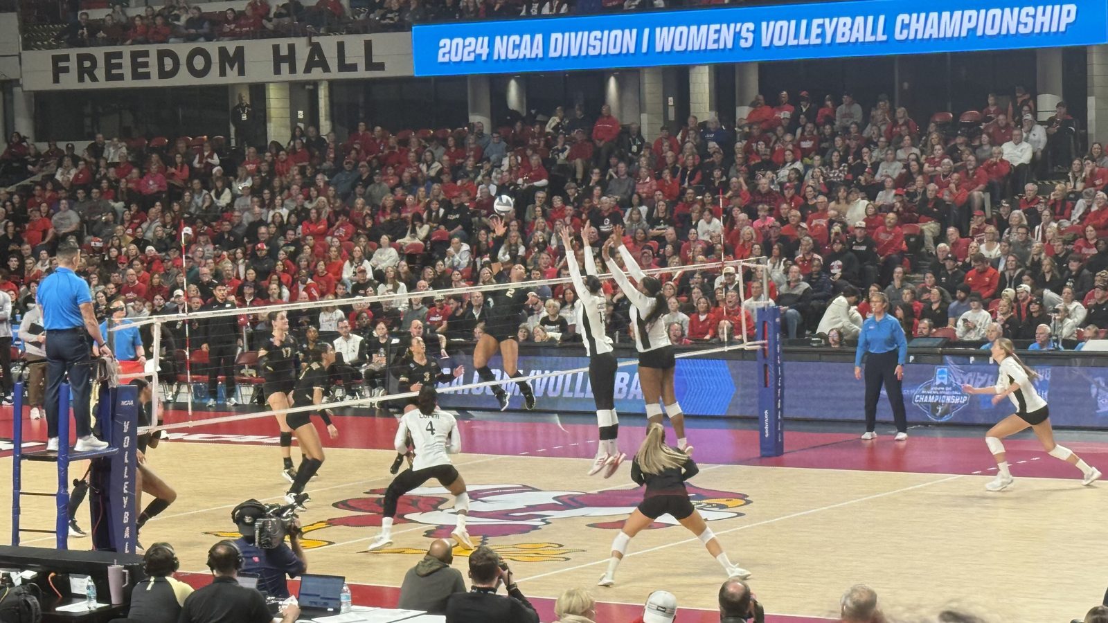 Purdue and Louisville women's volleyball players at the net during a 2024 NCAA Division I tournament match at Freedom Hall, with a packed crowd in red behind them.