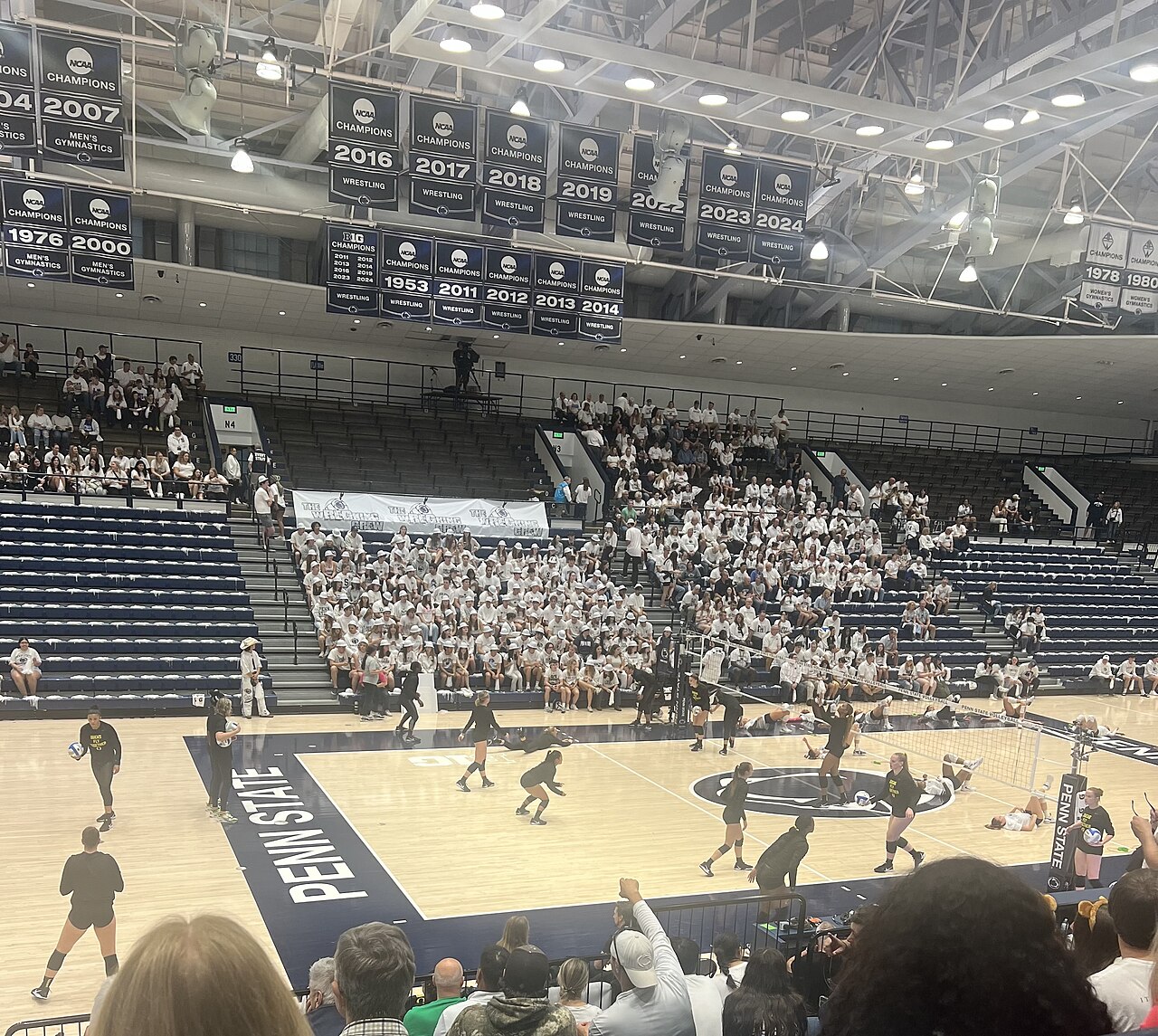 Penn State women's volleyball players in black warm-up shirts warm up on the Rec Hall court in front of a packed white-clad student section, with rows of championship banners hanging from the rafters.
