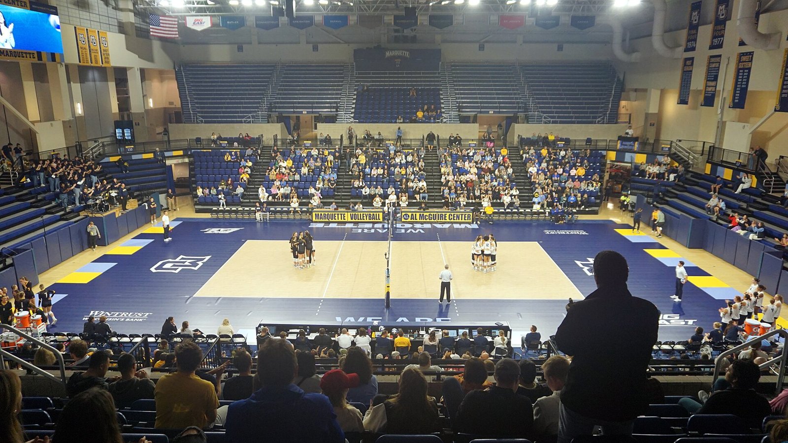 Wide view from the stands of a college volleyball arena with blue and gold seating, where two women's teams gather in separate huddles on either side of the net before the first serve.