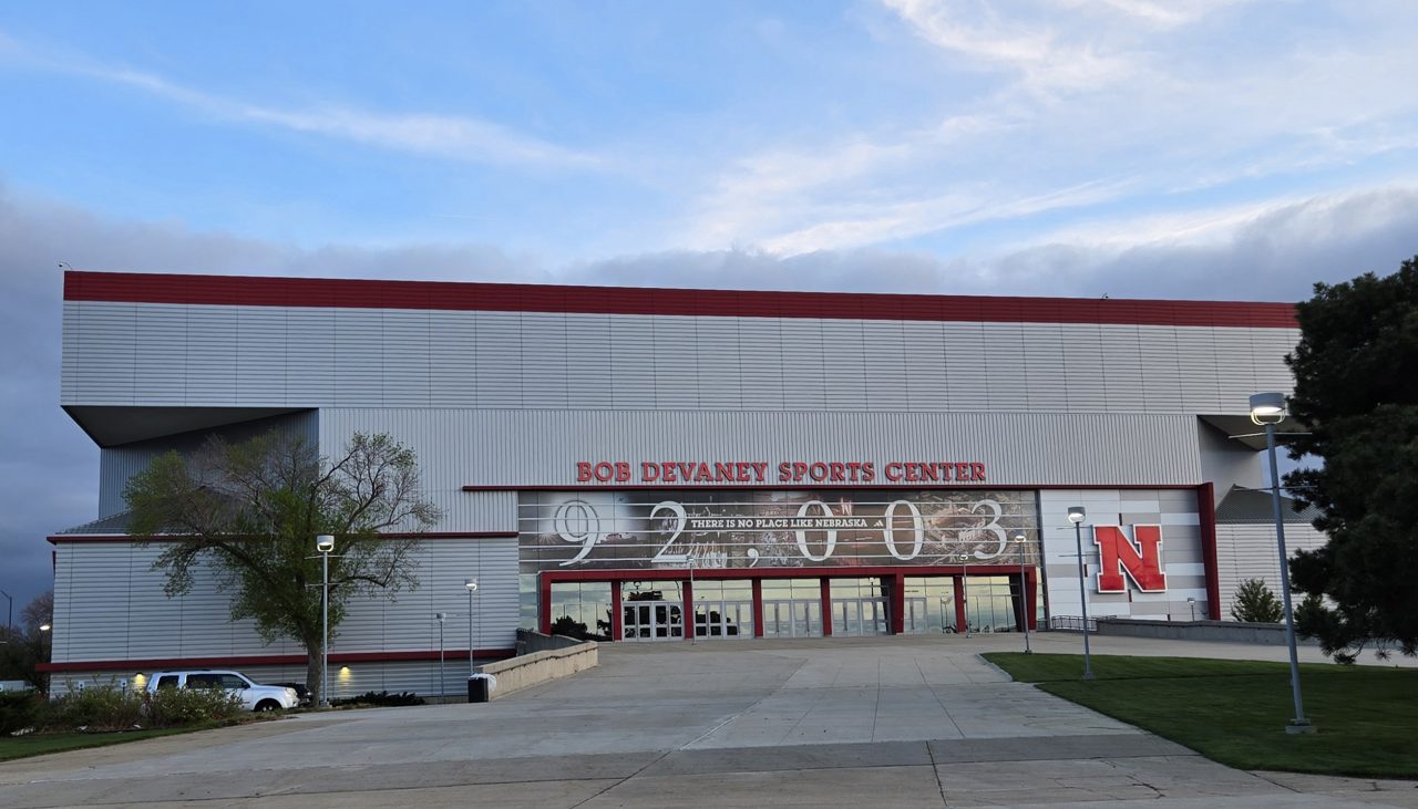 The north entrance of the Bob Devaney Sports Center in Lincoln, a long gray building with a red roofline, red block letters over the doors, a large 92,003 mural, and a red Nebraska N logo, under an evening sky.