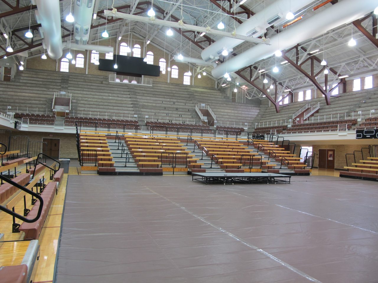 The interior of Gregory Gymnasium at the University of Texas, an older arena with arched windows, steel roof trusses, pale bleachers on three sides, and a covered floor, photographed empty in daylight.