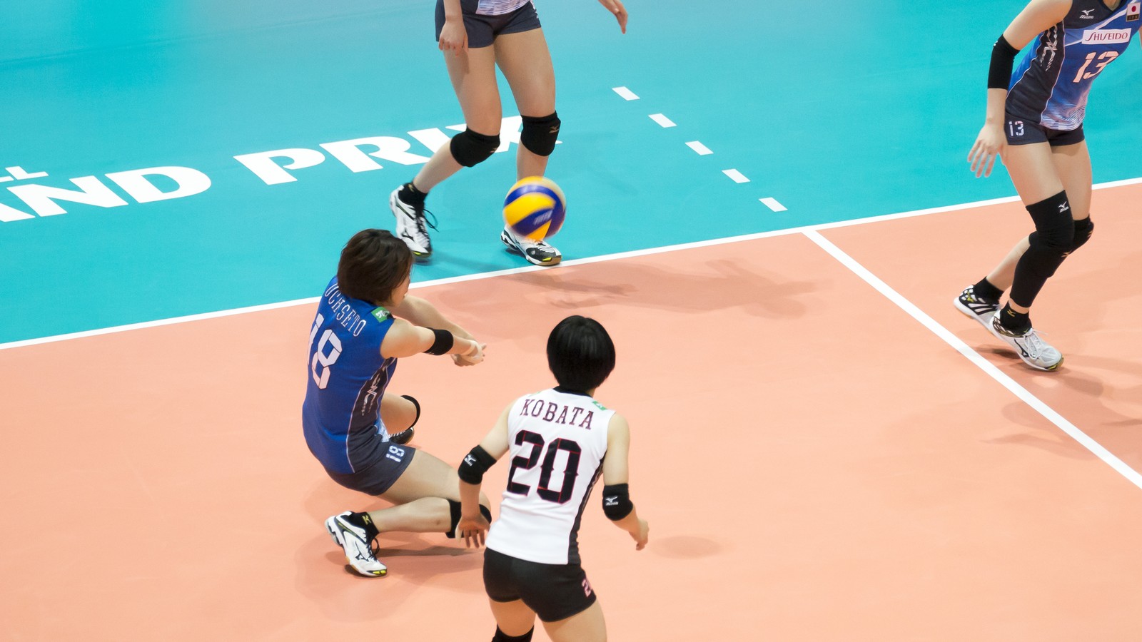 From above the court, a Japan women's national team player in a blue jersey number 18 lunges into a forearm pass while a teammate in a white libero jersey number 20 watches in a ready stance nearby.