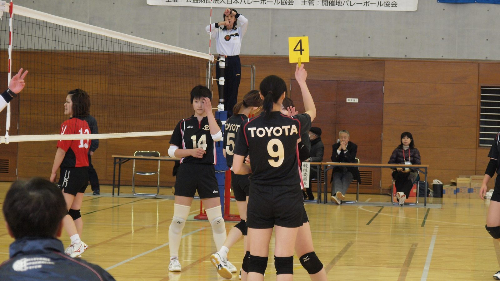 A women's indoor volleyball match in a wood-paneled gym: a player in a black Toyota jersey number 9 stands near the net while a teammate behind her holds up a yellow paddle printed with the number 4, and the first referee watches from the stand.