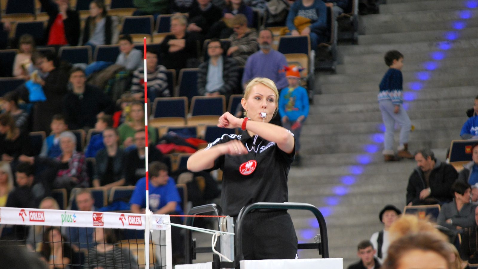 A referee with a whistle in her mouth stands on the referee stand at the net, rolling her forearms around each other in front of her chest, with spectators filling the stands behind her.
