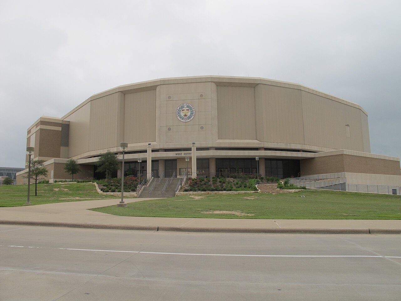 The tan, curved concrete exterior of Reed Arena at Texas A&M, with the university seal above the west entry, a wide staircase lined with landscaping, and a lawn and street in the foreground under a gray sky.
