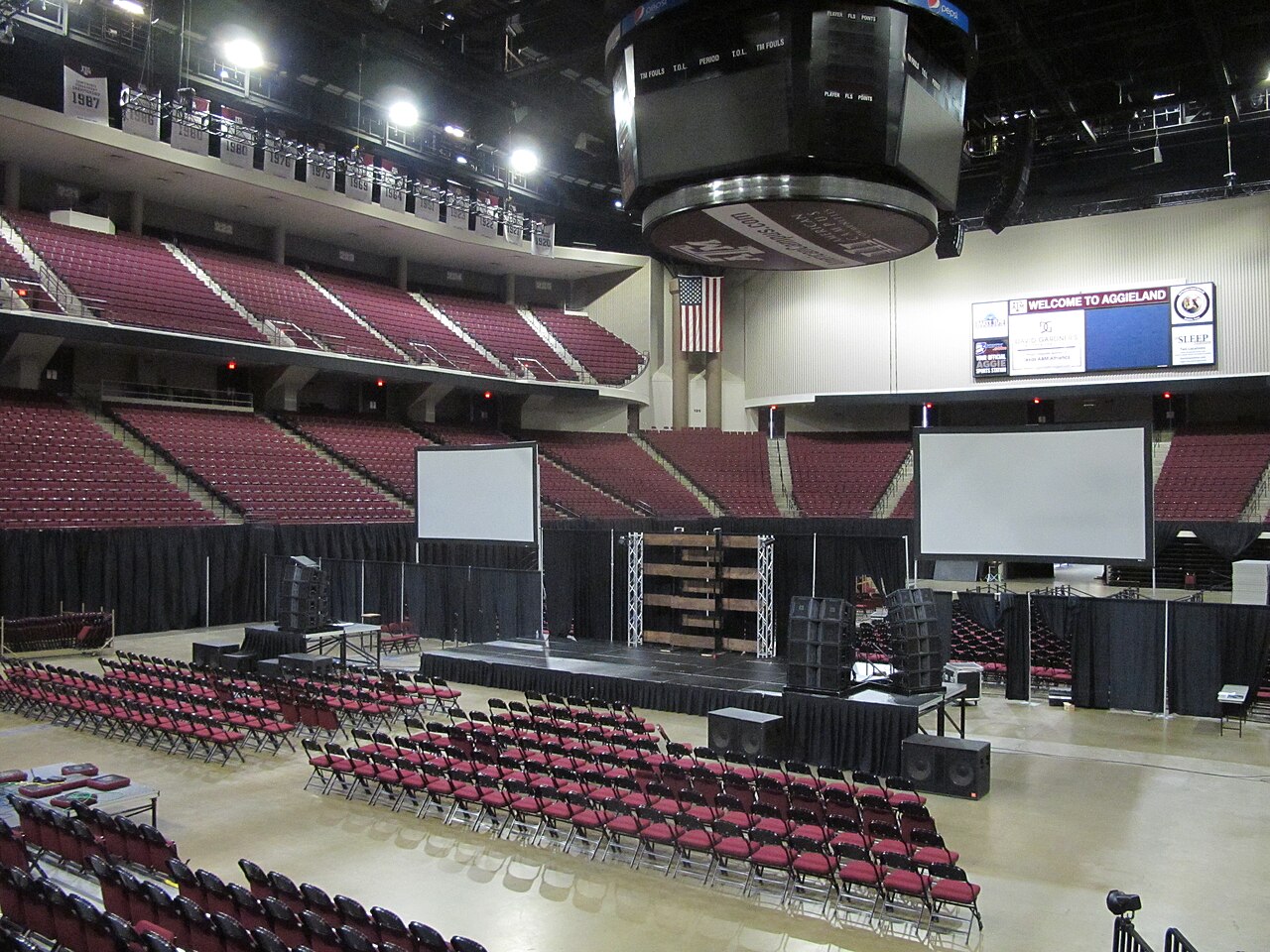The inside of Reed Arena with two tiers of maroon seats, a large center-hung scoreboard, and a floor set up with rows of red folding chairs and a stage for a campus event.