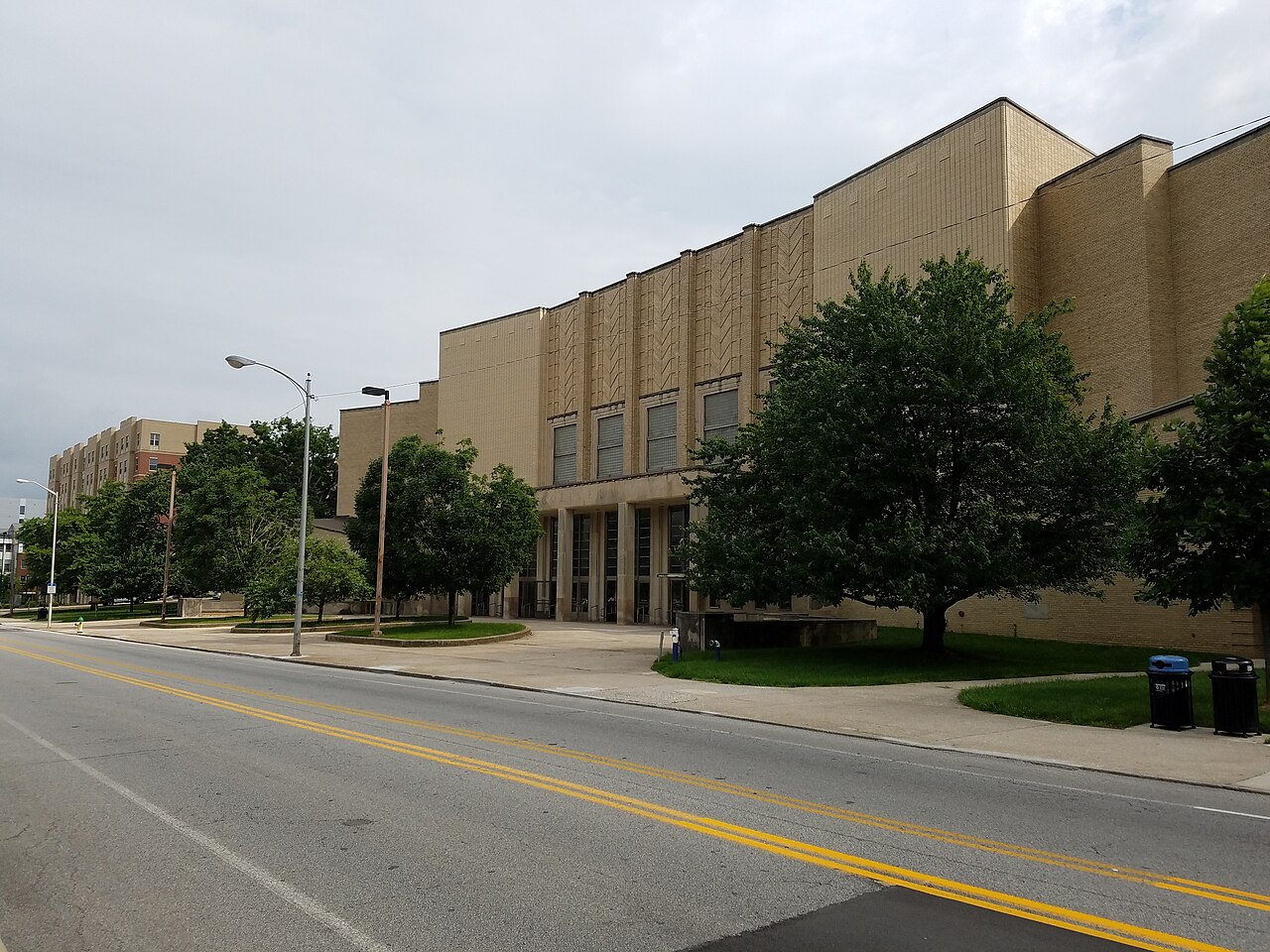 The tan brick, Art Moderne facade of Memorial Coliseum at the University of Kentucky, with tall vertical windows over a columned entrance, large shade trees on the lawn, and an empty street in the foreground under a cloudy sky.