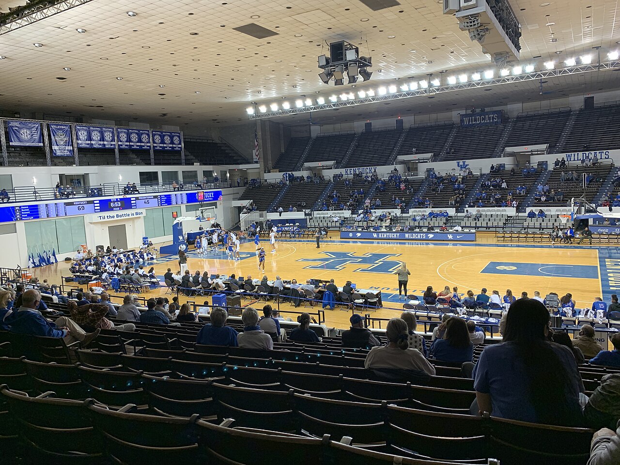 Inside Memorial Coliseum during a Kentucky women's basketball game, with blue seats in the foreground, fans spread through the lower bowl, a court with a large blue UK logo at center, and SEC championship banners hanging along the far wall.