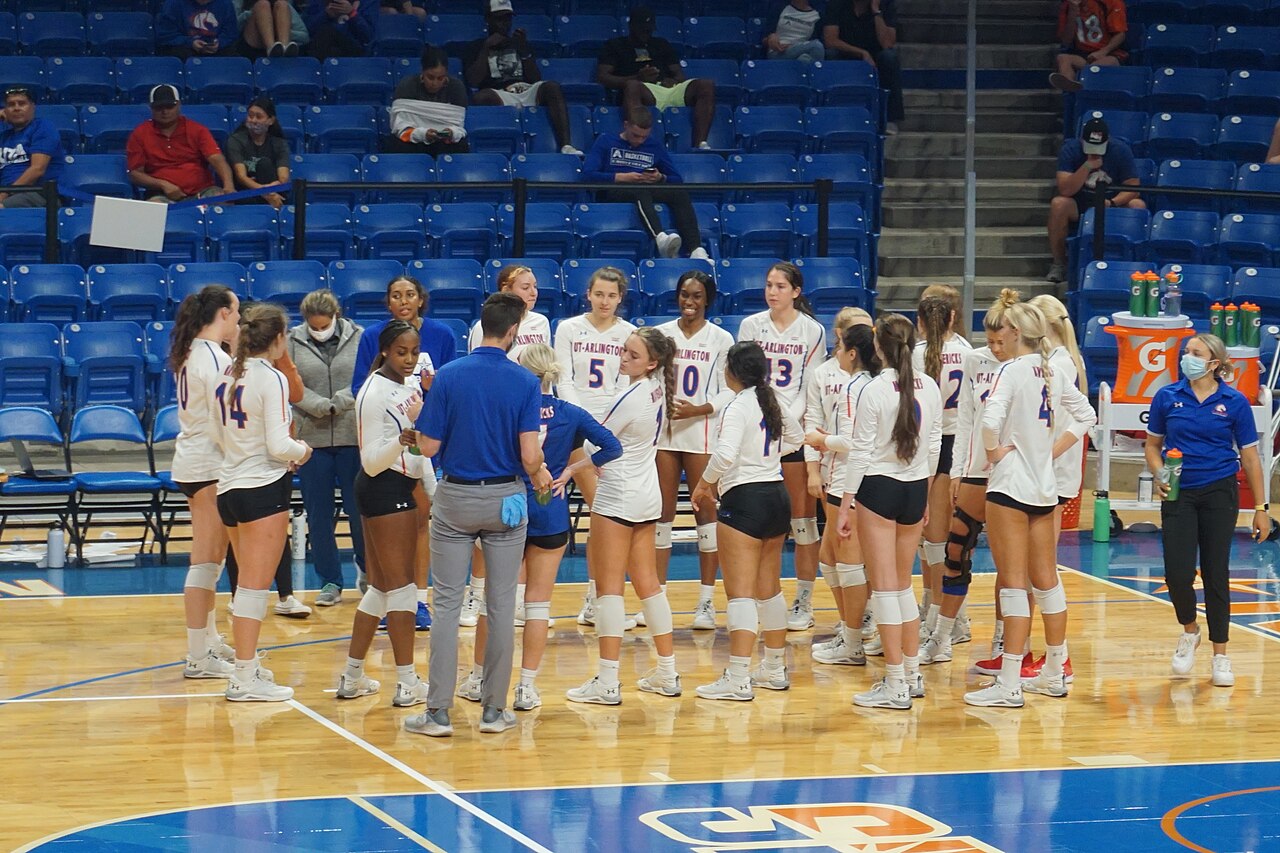 UT Arlington women's volleyball players in white jerseys and black shorts gather in a loose huddle on the court at College Park Center while a coach in a blue polo talks to them and a staff member stands near the Gatorade cart.