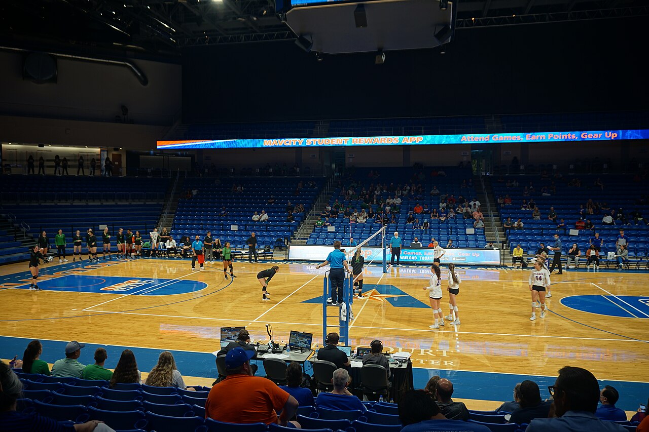 Wide view of College Park Center during a North Texas and UT Arlington women's volleyball match, with the full North Texas squad standing along the far sideline in black and green while players on the court wait for a serve and fans sit in blue seats.